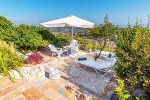 Eine Terrasse im Freien mit Steinböden, weißen Lounge-Stühlen, einem weißen Sonnenschirm und Topfpflanzen.