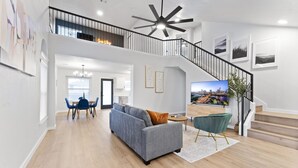 A spacious living area with a grey sofa, a dining table with chairs, a staircase, and a cityscape TV.