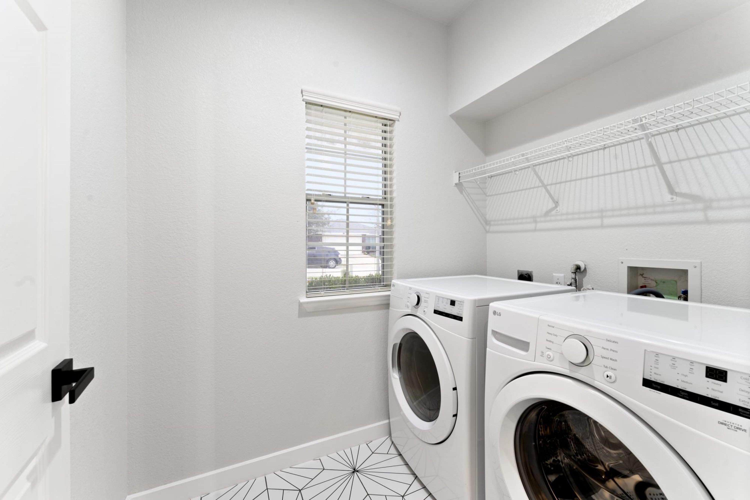 A laundry room with two white front-loading washing machines, a window with blinds, and a metal drying rack.