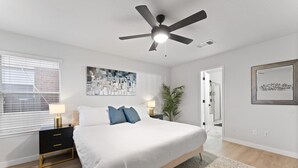 A bedroom with a bed, a ceiling fan, a nightstand with a lamp, a wall art piece, and a window with blinds.