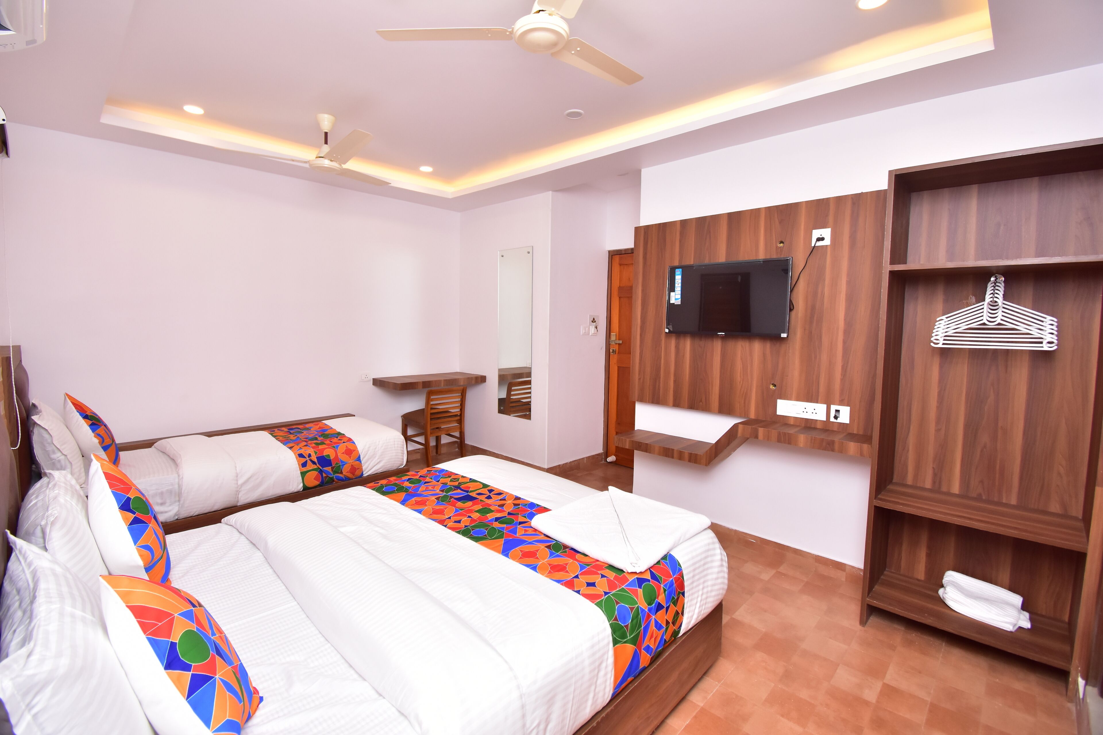 A hotel room with two beds, a TV mounted on the wall, a wooden wardrobe, and a desk with a chair.