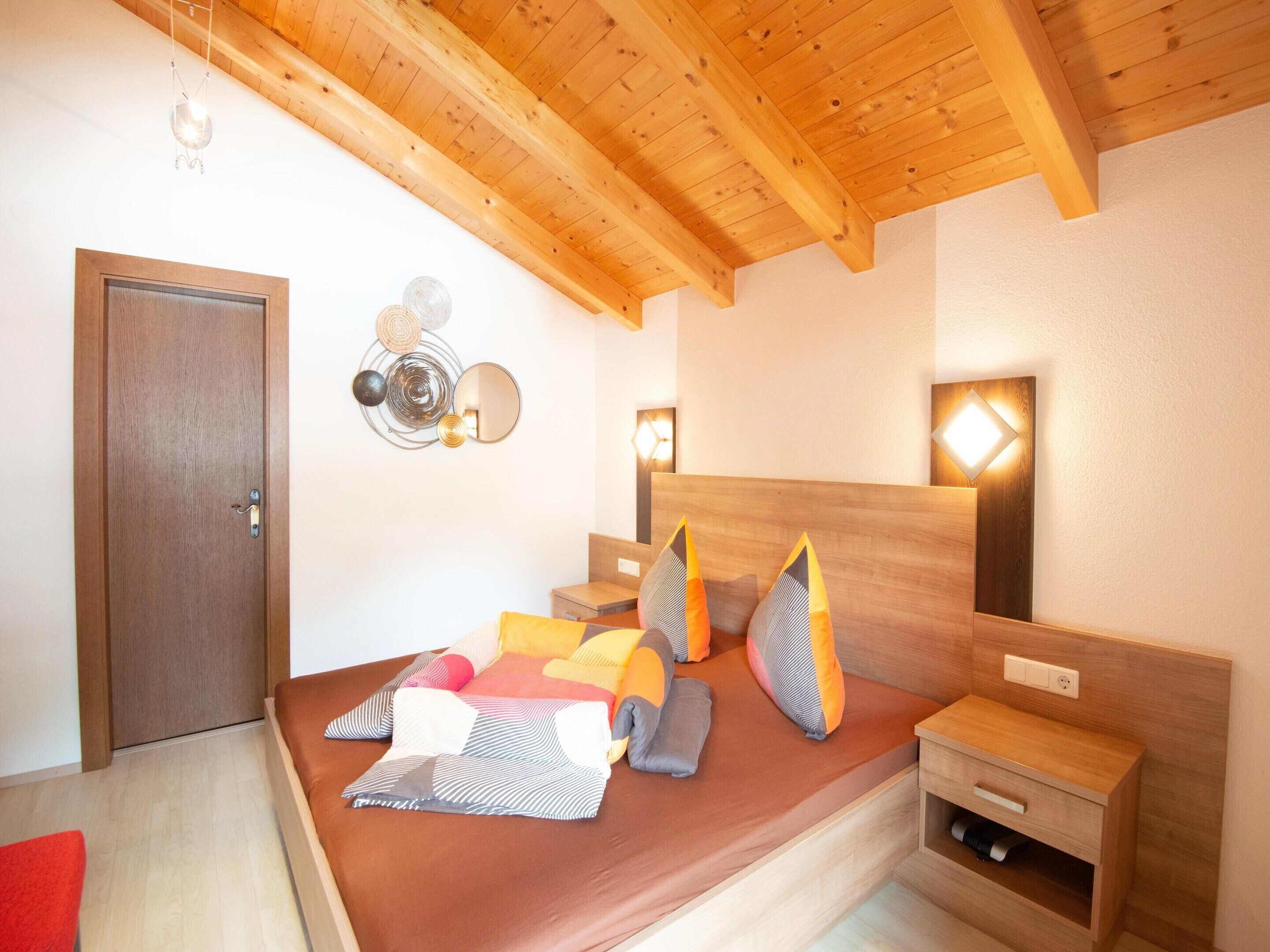 A bedroom with a wooden ceiling, a bed with colorful pillows, a bedside table, and a wall-mounted light fixture.