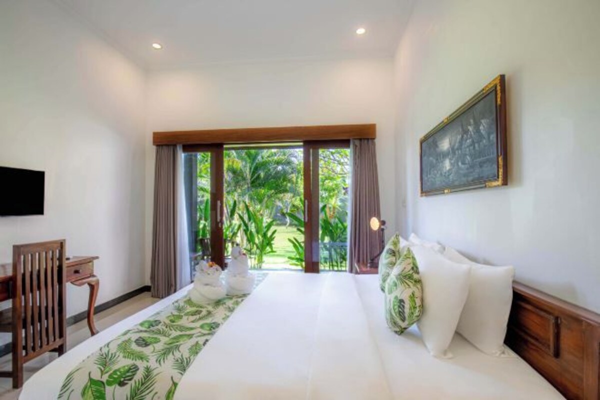 A bedroom with a large bed, a desk, and a view of greenery through the window.