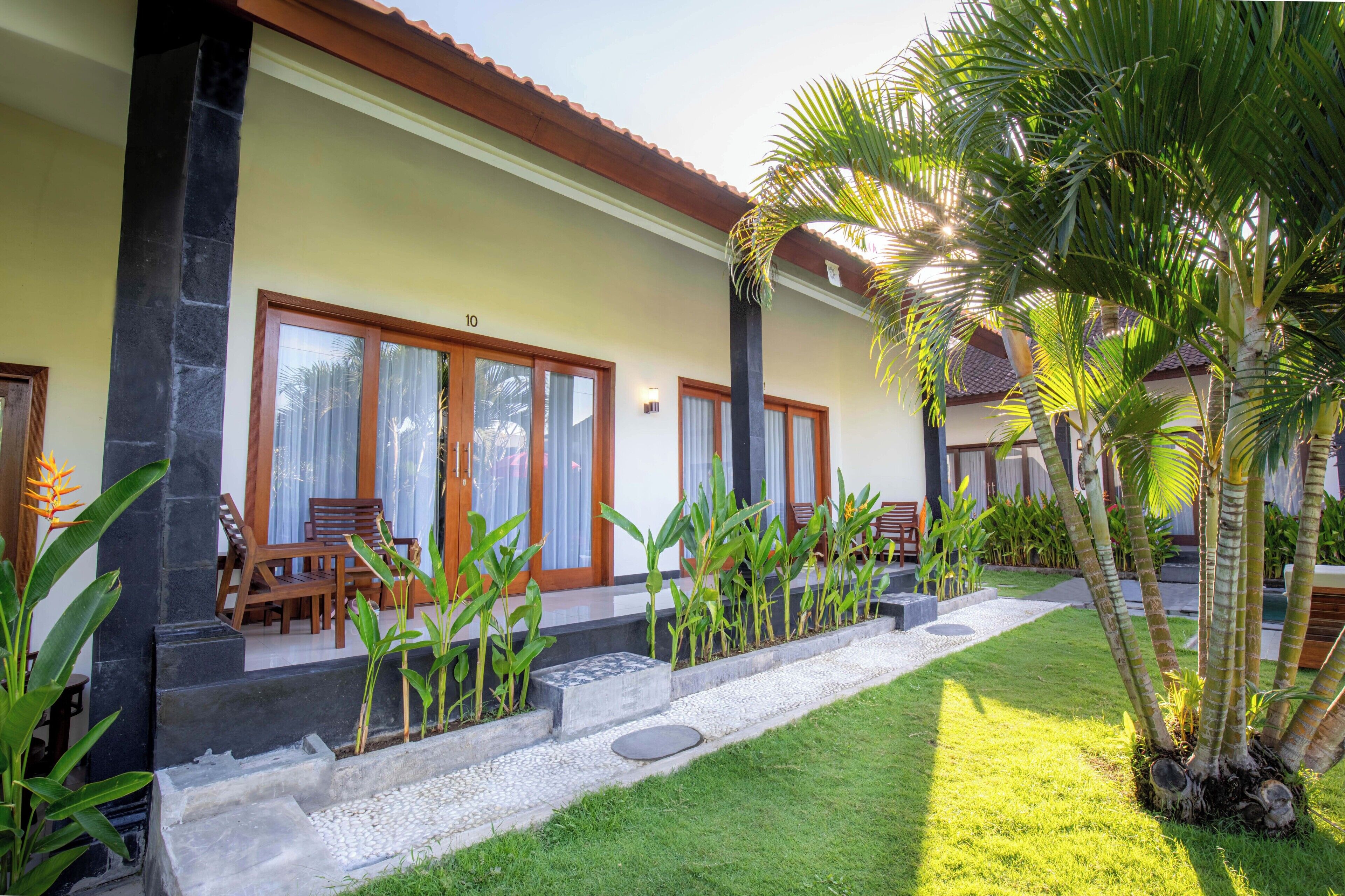 A building with large windows, a stone pillar, a wooden bench, and a well-maintained lawn with palm trees.