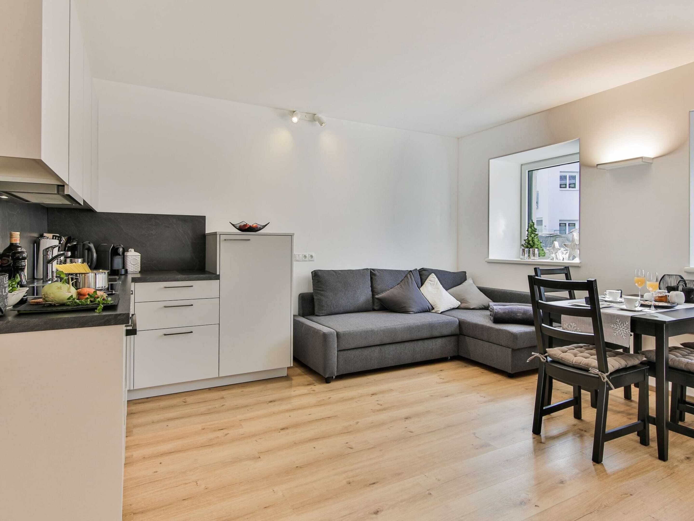 A modern kitchen with a grey sofa and dining table, wooden flooring, and a window with a city view.
