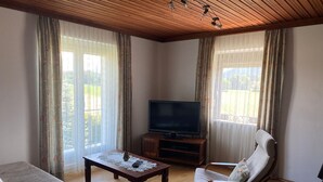 Uma sala de estar com teto de madeira, uma televisão sobre um suporte de madeira, um sofá, uma poltrona e uma mesa de centro com um controle remoto e um objeto decorativo.