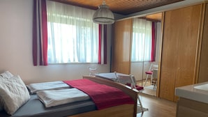 Quarto com cama, escrivaninha e cadeira. Cortinas nas janelas e um guarda-roupa visível ao fundo.