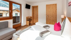 A bedroom with a bed, a grey sofa, a wooden desk, a TV, and a window with a view of buildings and mountains.