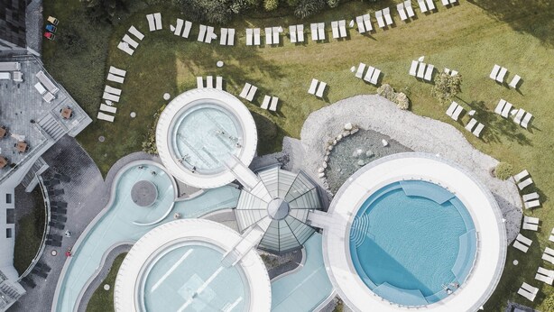 Vista aérea de un moderno complejo turístico con múltiples piscinas circulares, tumbonas y una fuente central.