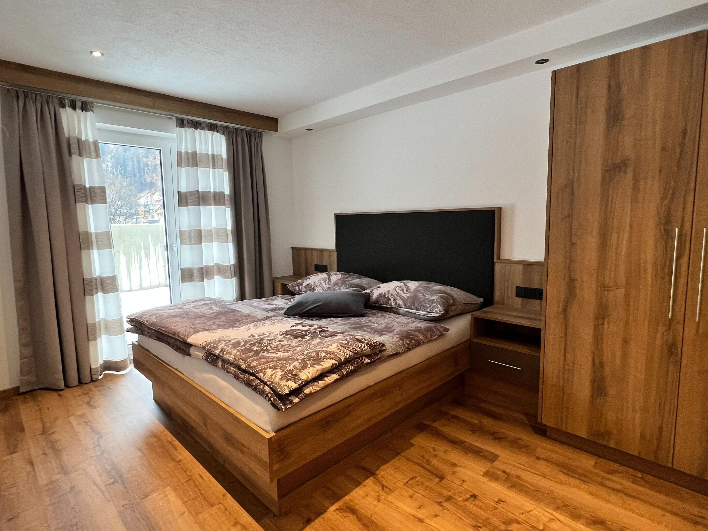 A modern bedroom with a wooden bed, a large wardrobe, and a view of the outdoors through a window with curtains.