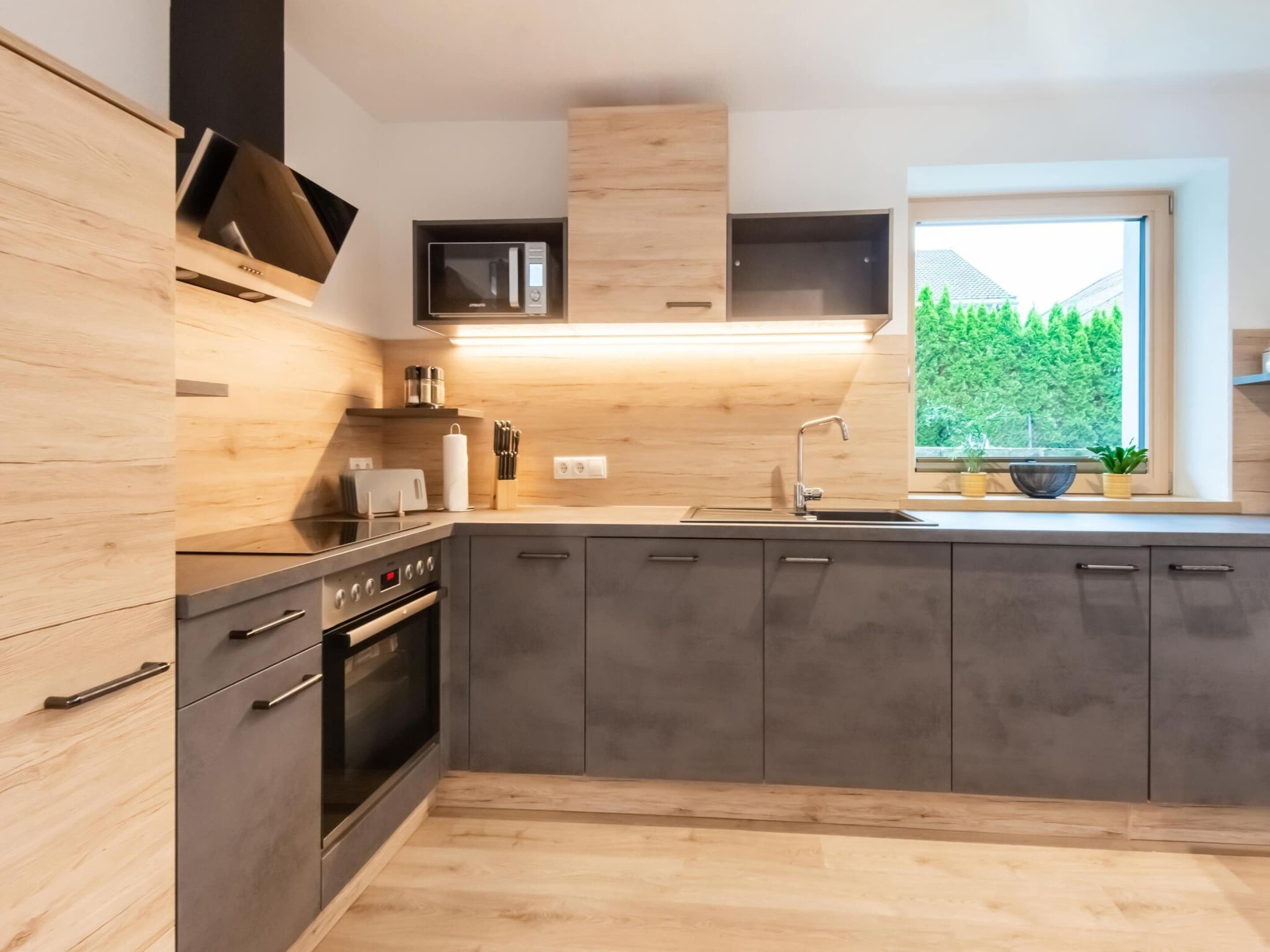 A modern kitchen with dark cabinets, a built-in oven, a microwave, and a window with a view of greenery.