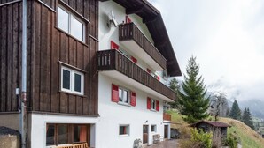 Un immeuble à plusieurs étages, avec une façade en bois et des volets rouges, un banc en pierre à l’extérieur, et une chaîne de montagnes en arrière-plan.