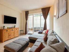 A living room with a sofa, ottoman, wooden cabinet, TV, and a balcony with curtains. - Apartment Zwieselstein Near Ski Slopes (Soelden)