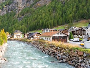 Un fiume con un litorale roccioso, una fila di case di legno con tetti spioventi e un'imponente montagna ricoperta di boschi sullo sfondo.
