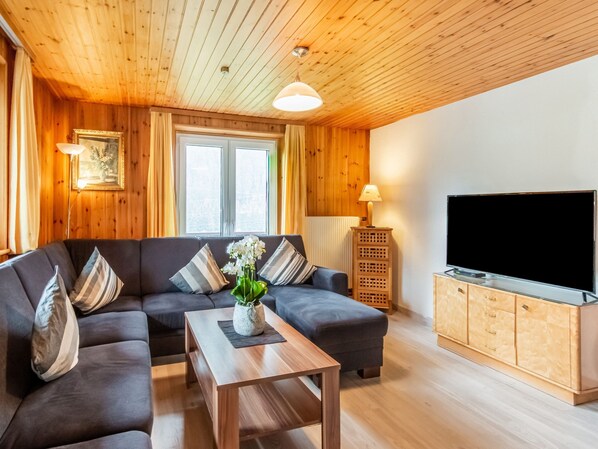 Un salon moderne avec un plafond en bois, un canapé d’angle gris, une table basse en bois, un téléviseur posé sur un meuble en bois et une fenêtre avec des rideaux.