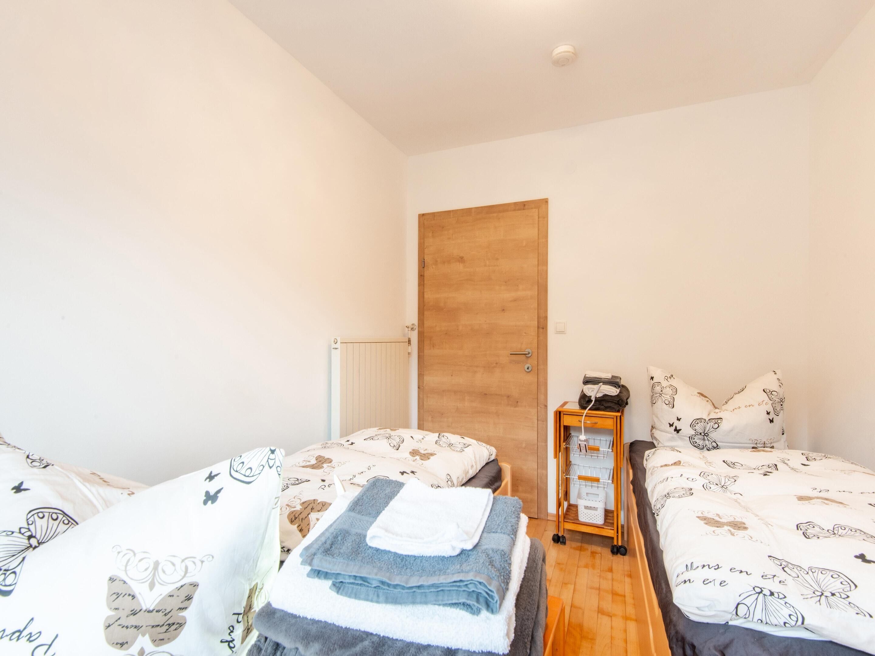 Une petite pièce bien éclairée, avec deux lits, une porte en bois, un radiateur et une petite table d’appoint en bois sur laquelle se trouve un panier.