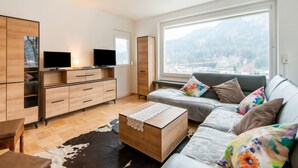 A modern living room with a sofa, coffee table, TV, and a large window with a mountain view. - Holiday Home Feldkirch Panoramic Views (Feldkirch)