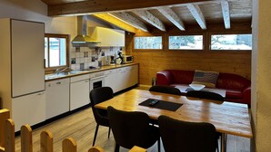 A compact kitchen with wooden beams, a dining area with a red sofa, and a view of trees through the windows. - Apartment in Montafon Near Ski Lifts (Sankt Gallenkirch)