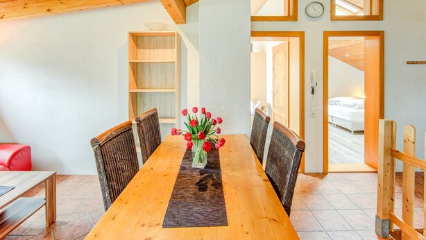 A dining area with a wooden table, wicker chairs, and a vase of red flowers. There is a wooden bookshelf and a door leading to another room. - Apartment in Montafon Near Ski Lifts (Sankt Gallenkirch)