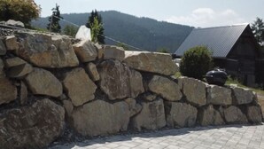 A stone wall with a paved area in front, a wooden cabin with a metal roof, and a mountainous background. - Apartment in Salchau Near Lachtal Ski Area (Oberwölz)