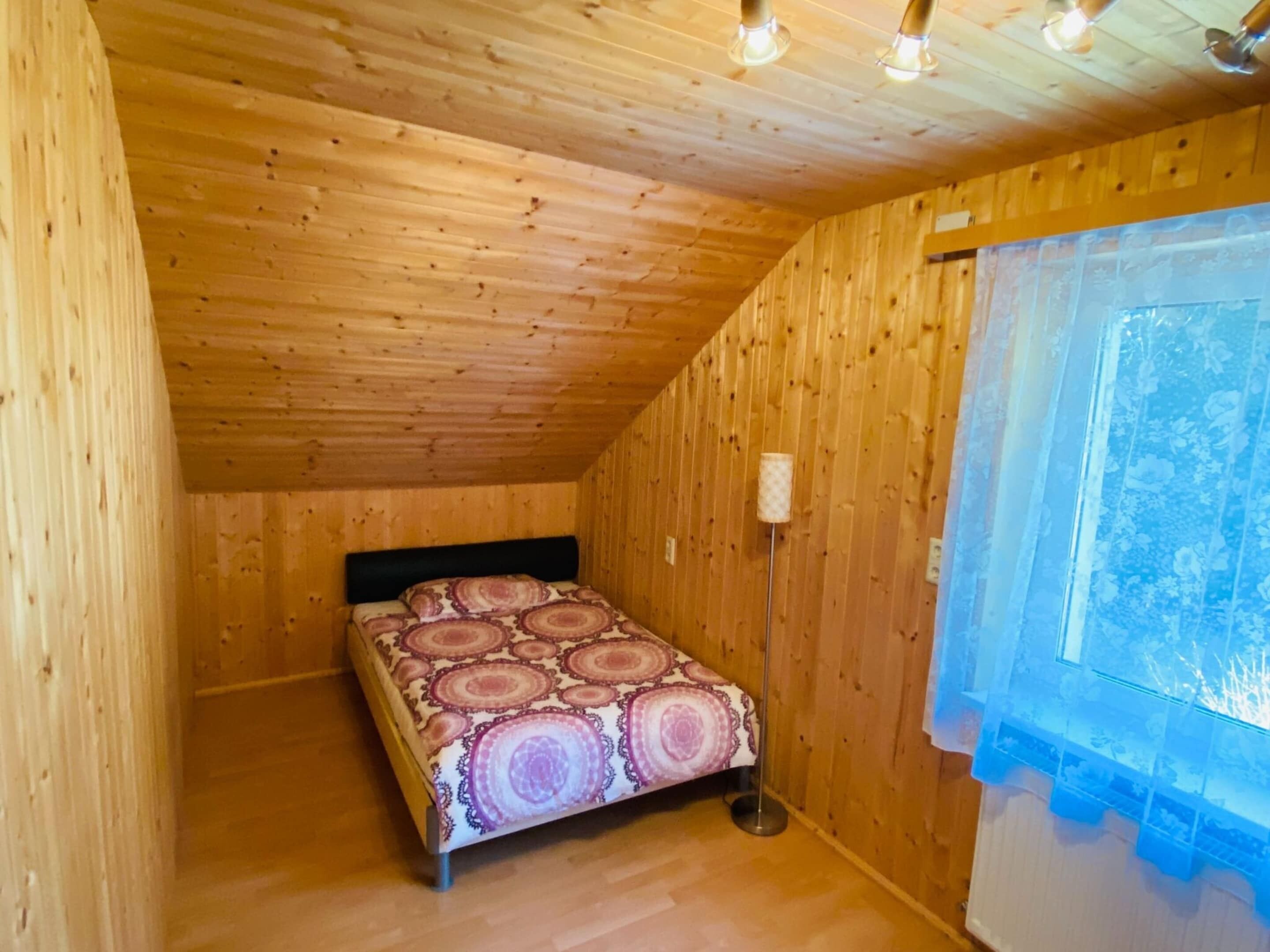 Um quarto pequeno e acolhedor com paredes e teto de madeira, uma cama com colcha estampada, um abajur e uma janela com vista para vegetação.