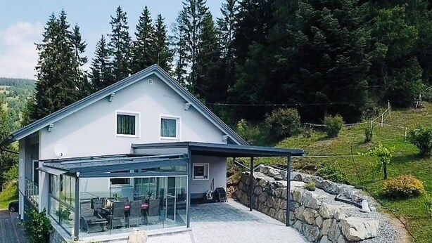 A modern house with a glass-enclosed patio area, stone retaining walls, and a view of a forested hillside. - Apartment in Salchau Near Lachtal Ski Area (Oberwölz)