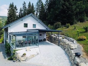 Una casa moderna con un patio cubierto de vidrio, muros de contención de piedra y vistas a una ladera boscosa.