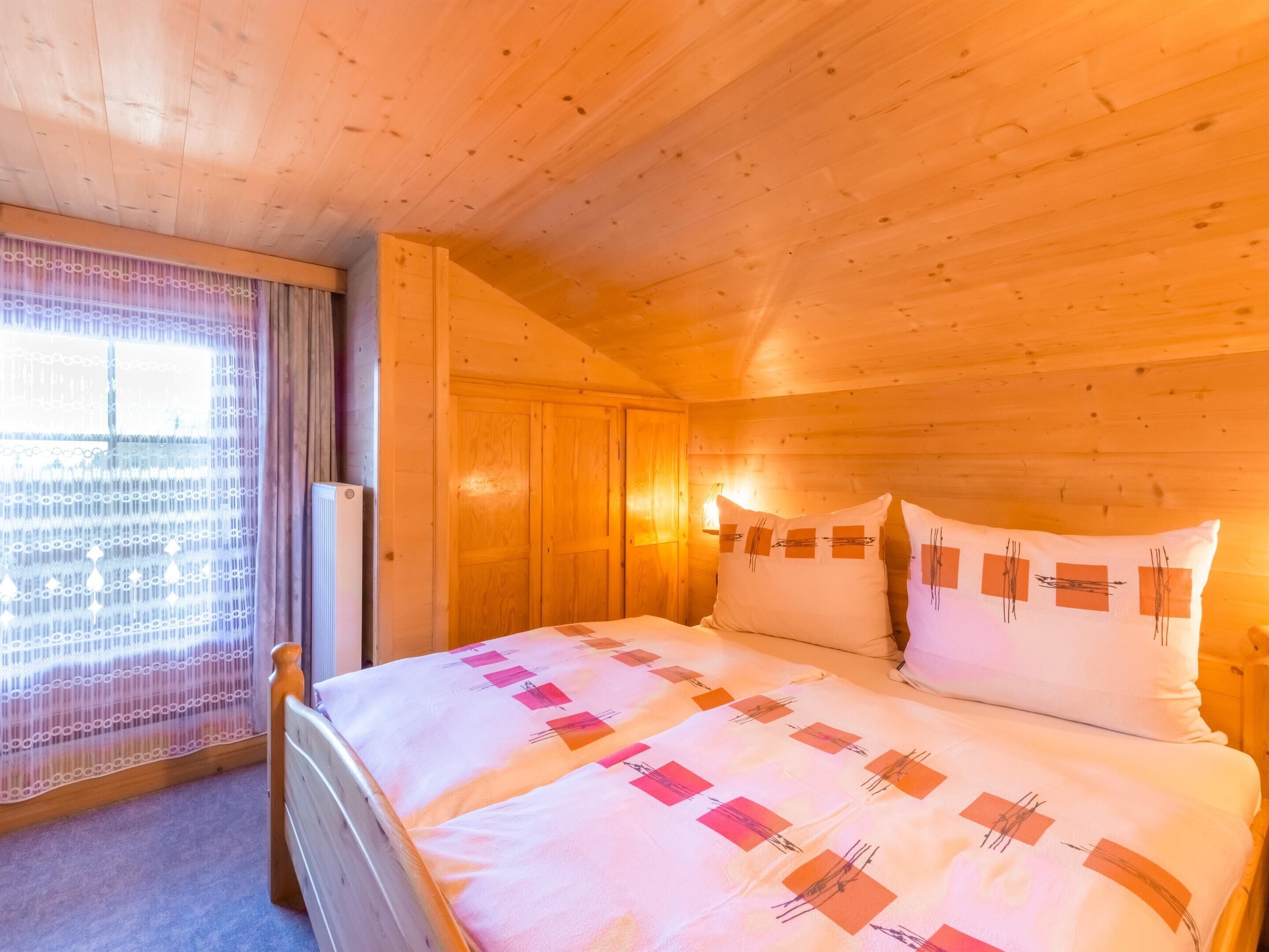 Ein Zimmer in einer Holzhütte mit einem Bett, einem Fenster mit Vorhang und einer Wandleuchte.