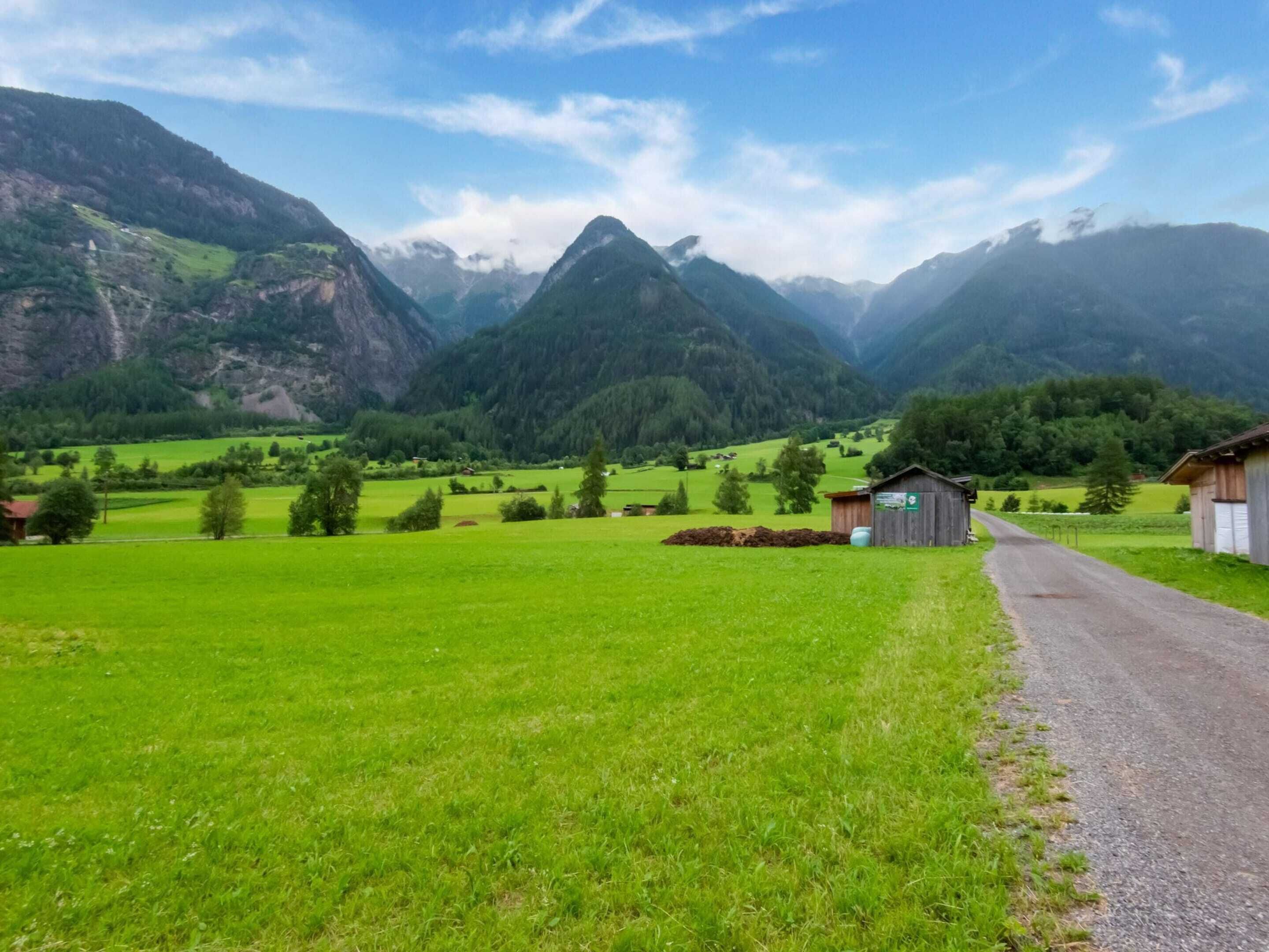 A rural landscape with a dirt road leading to a wooden building, surrounded by green fields and mountains.