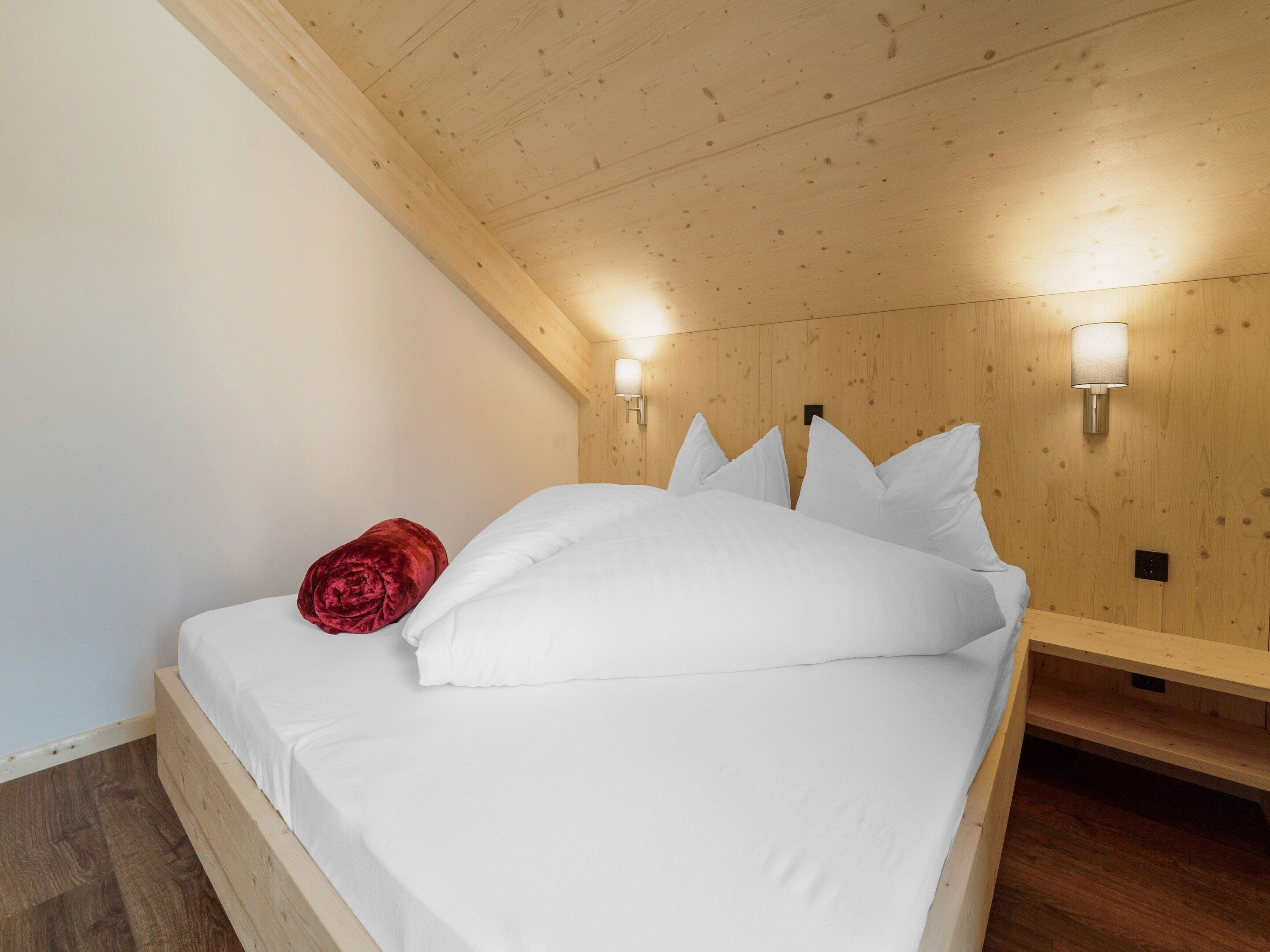 A wooden ceiling with a sloped design, a bed with white bedding and a red pillow, and wall-mounted lighting.