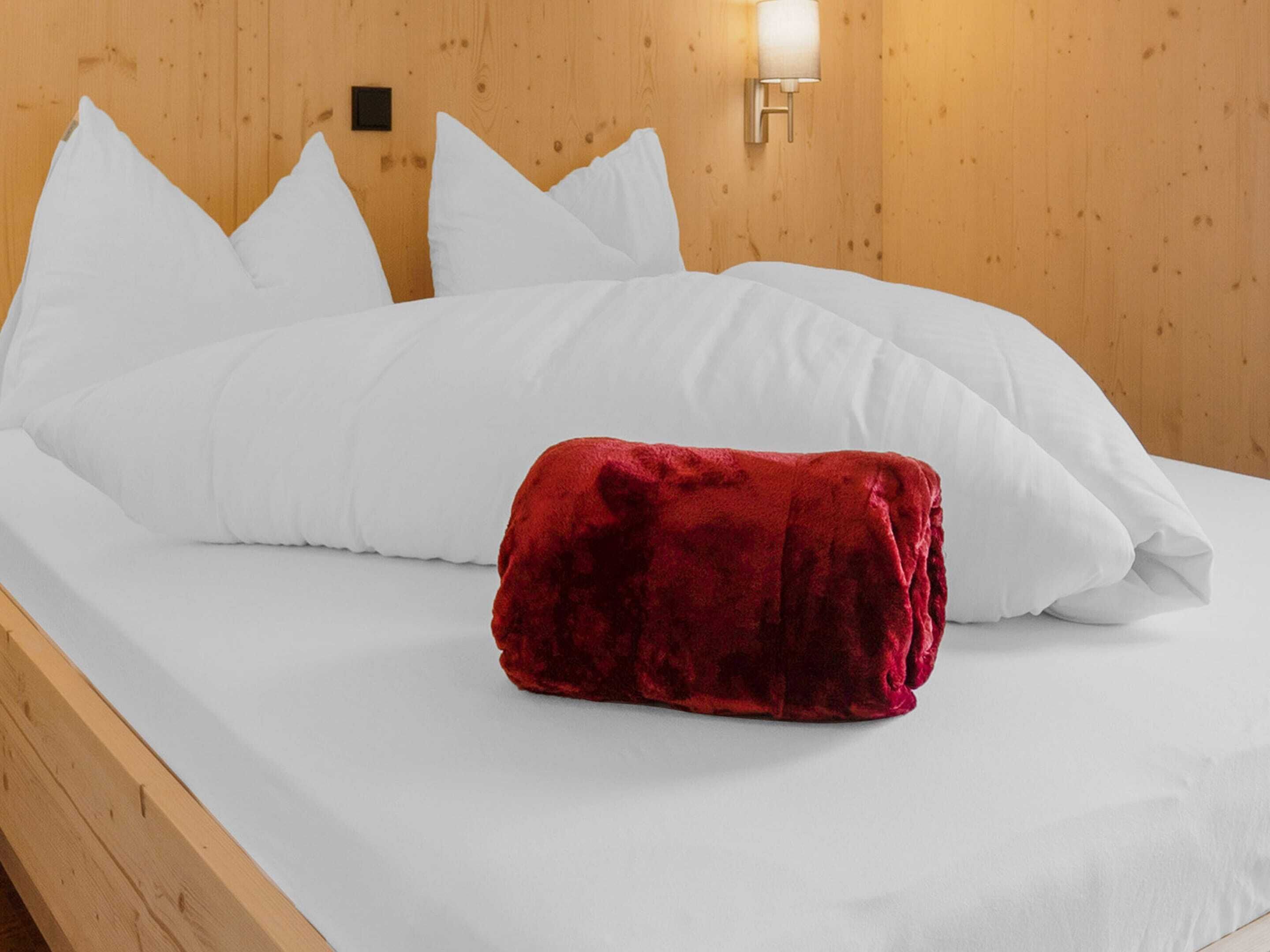 Un lit avec des oreillers blancs et un oreiller décoratif rouge, une tête de lit en bois et un luminaire fixé au mur.