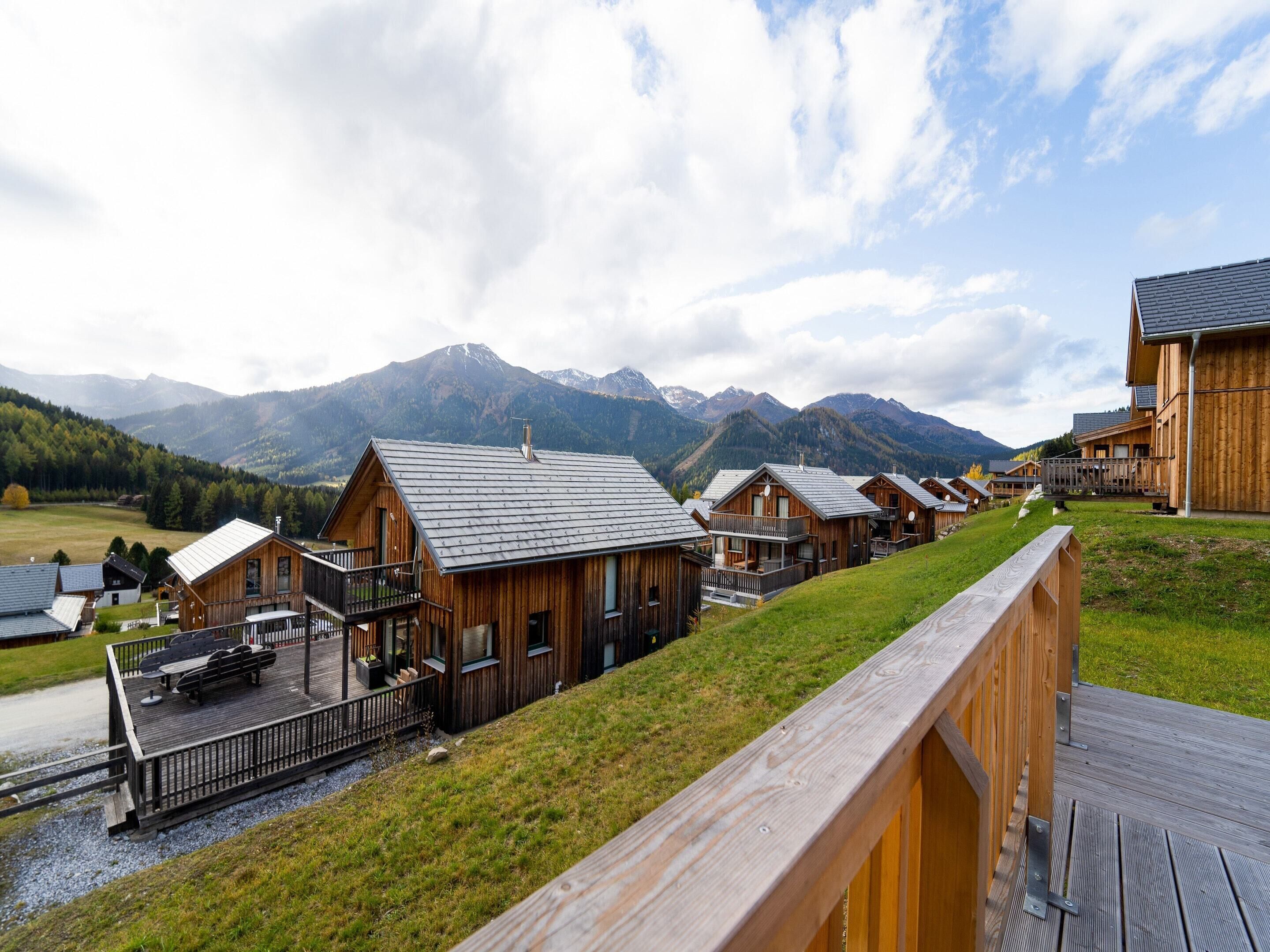 A row of wooden cabins with balconies, set against a backdrop of mountains and a cloudy sky.