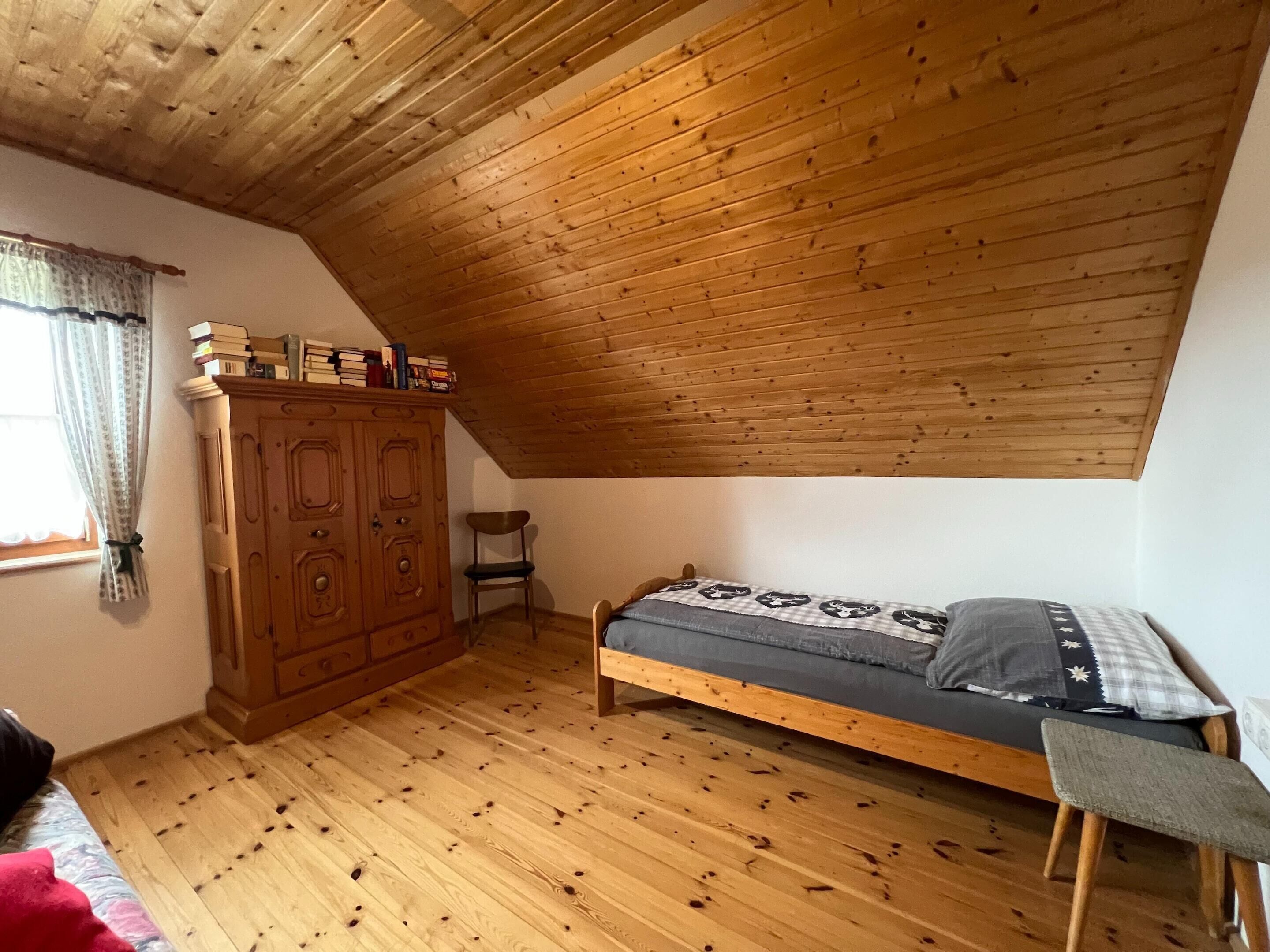 A small, cozy room with a sloped ceiling, wooden floor, a bed, a wooden wardrobe, and a window with curtains.