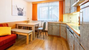 A compact kitchen with a dining area, wooden furniture, and a view through a window with orange curtains. - Apartment in Serfaus-fiss-ladis (Ladis)