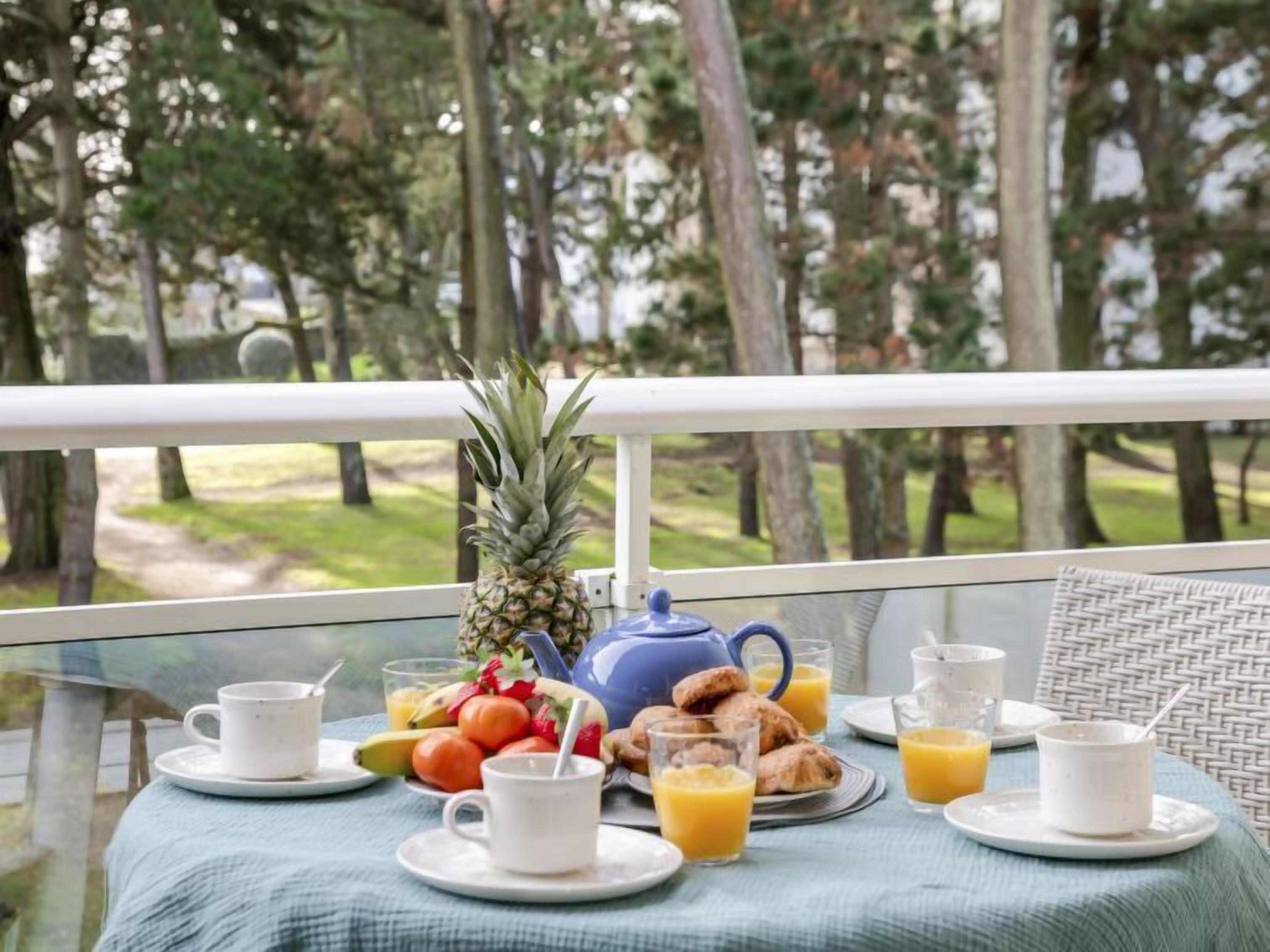 A breakfast table with a pineapple, fruit, croissants, and orange juice on a balcony with a view of trees.