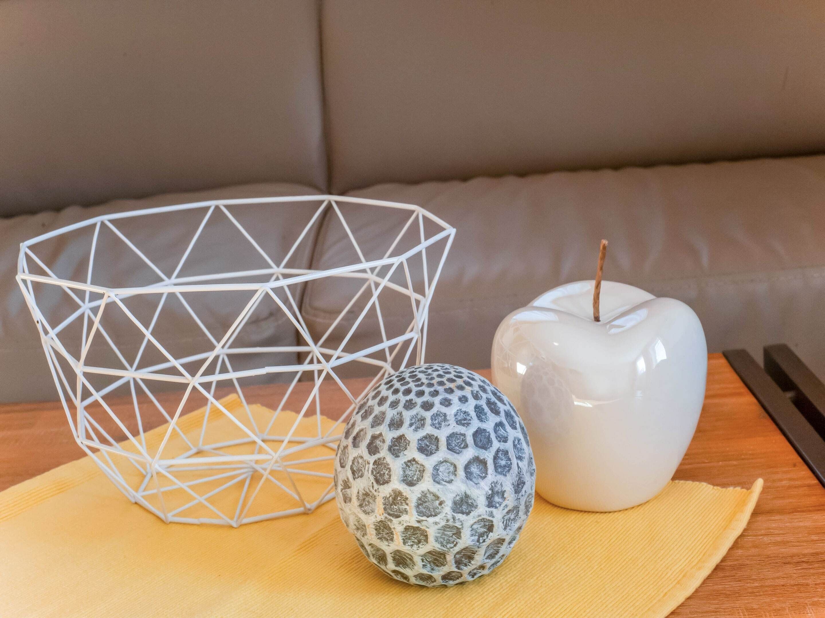 A white geometric wire basket, a patterned spherical object, and a white apple with a stick on a wooden surface.