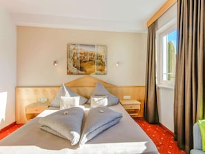 A hotel room with a large bed, two bedside tables, a nightstand, a painting on the wall, and a window with curtains.