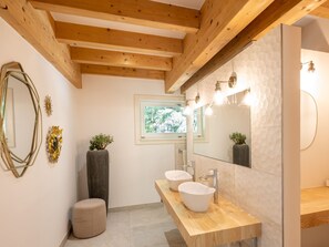 Une salle de bain moderne avec des poutres en bois, un grand miroir, deux vasques et une fenêtre donnant sur un paysage verdoyant.