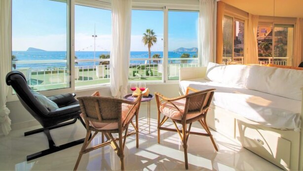 Ein Hotelzimmer mit Balkon, einem weißen Sofa, zwei Korbstühlen, einem kleinen Tisch mit Flasche und Gläsern sowie Blick auf das Meer und die Berge.