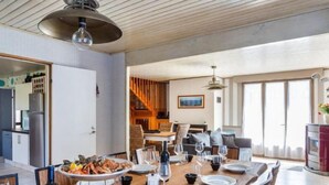 A dining area with a wooden table set for a meal, a kitchen in the background, and a view of the outdoors through large windows. - La Perle by Interhome (Cancale)