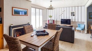 A modern living room with a wooden dining table, wicker chairs, a grey sofa, and a flat-screen TV. - La Perle by Interhome (Cancale)