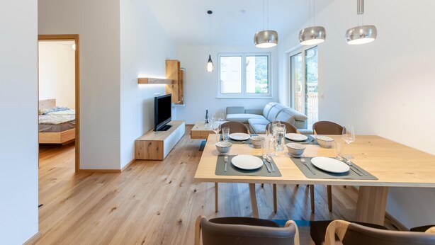 Un espace de repas moderne avec une table en bois dressée pour quatre personnes, un téléviseur et un canapé en arrière-plan.