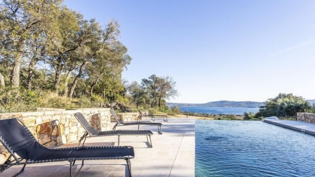 Un espace aménagé au bord de la piscine, avec des chaises longues et un mur en pierre, offrant une vue sur une étendue d’eau et des collines lointaines.