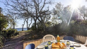 Une table à manger installée à l’extérieur, avec des assiettes, des tasses et un panier de pain, offrant une vue panoramique sur un paysage magnifique, avec des arbres et la mer à l’horizon.