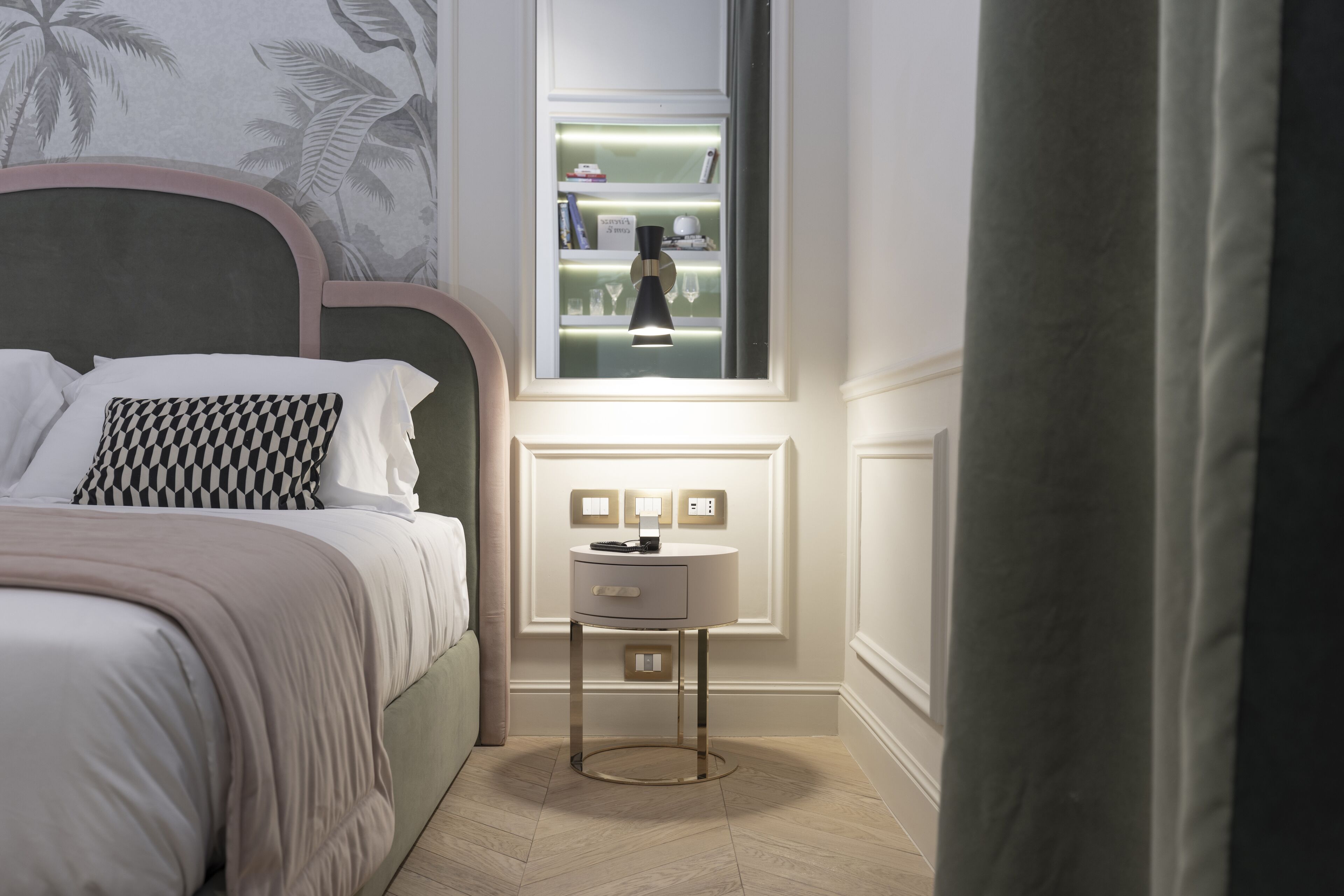 A bedroom with a bed, a bedside table with a lamp, a window with shelves, and a wall with a leaf pattern.