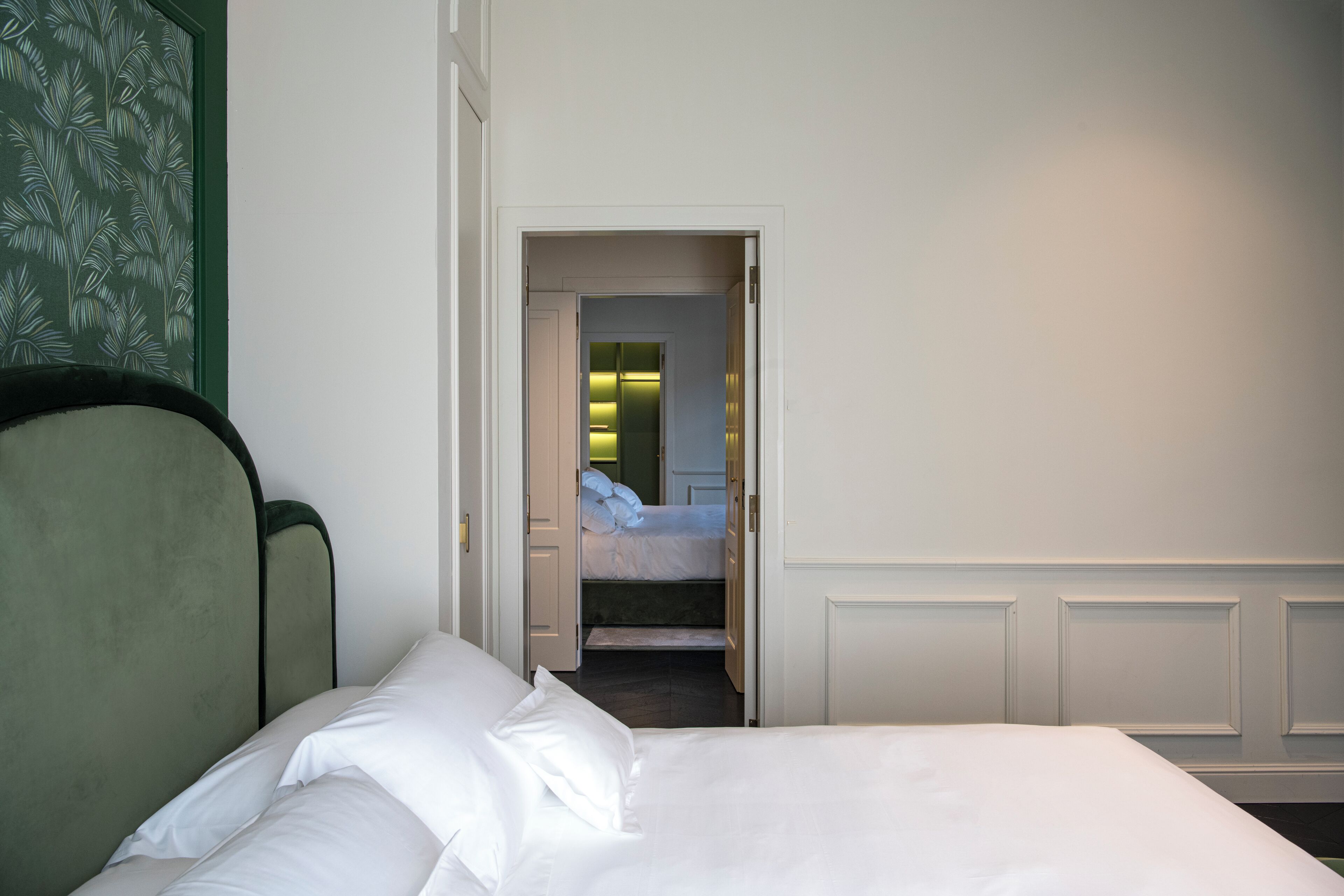 Habitación de hotel con cama, cabecero tapizado de color verde y puerta que da a un baño con espejo e iluminación.
