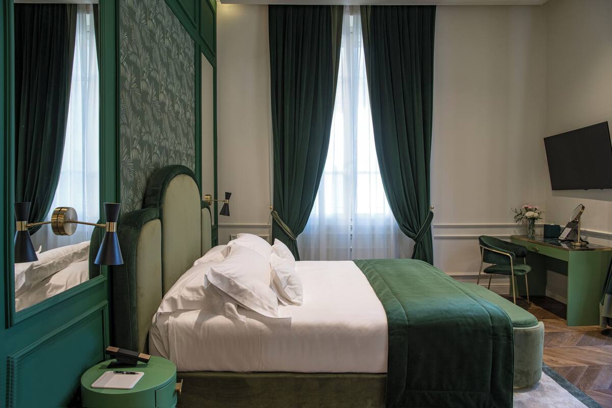 A hotel room with a bed, a desk, a chair, a flat-screen TV, and a window with curtains.