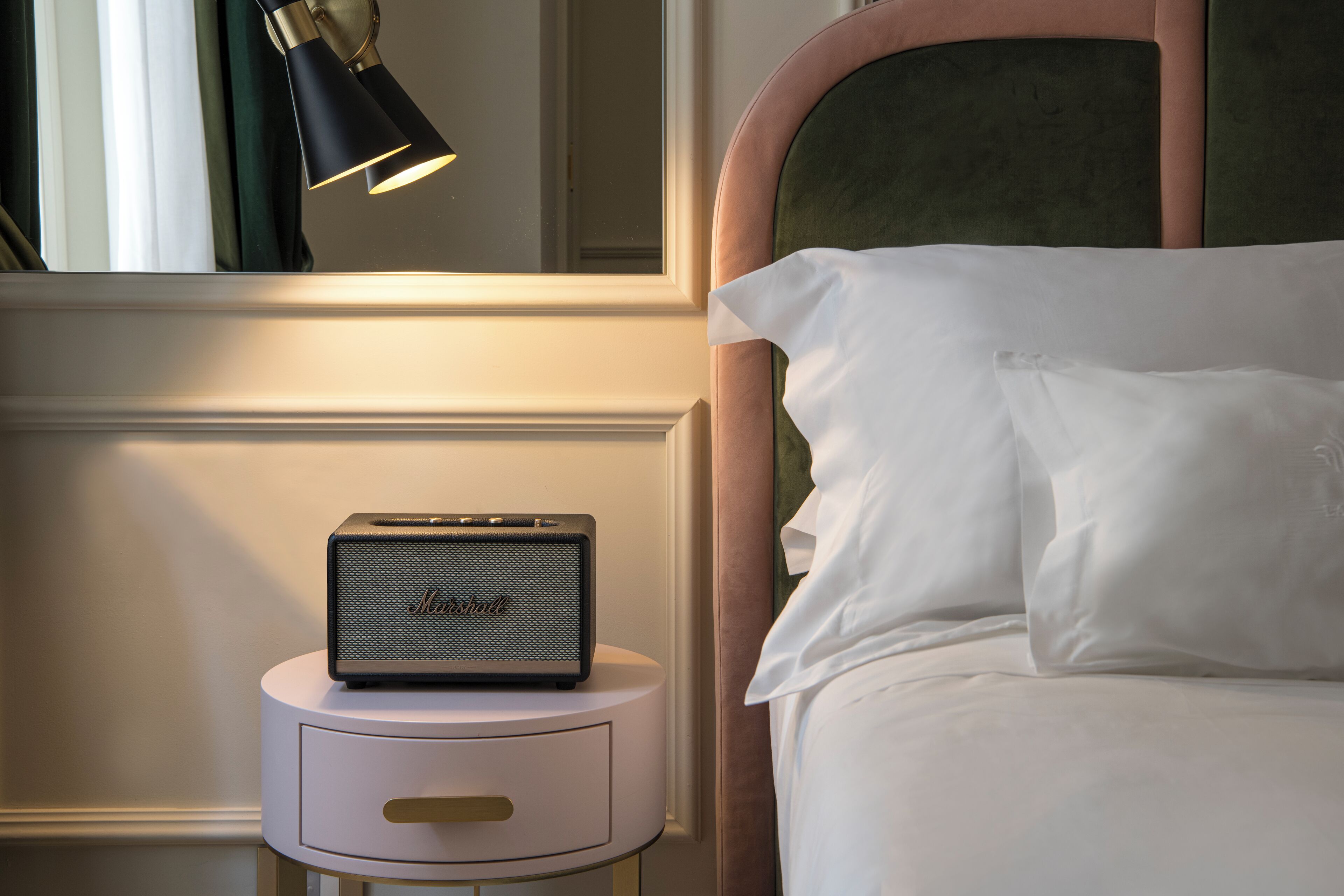 A neatly made bed with white linens, a small bedside table with a lamp, and a Marshall speaker.
