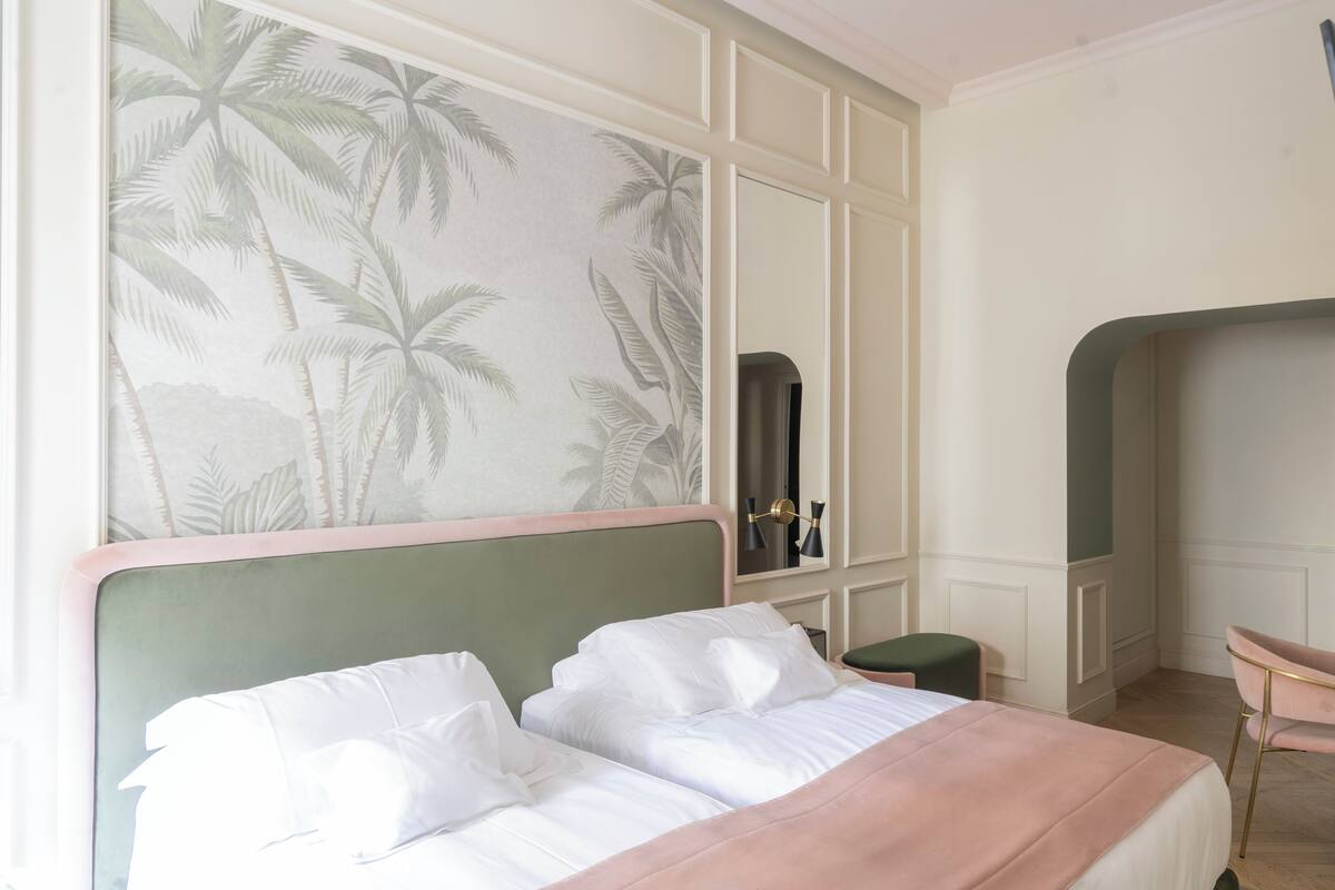 A bedroom with a large bed, a green headboard, a wall with a palm tree wallpaper, a mirror, and a small green stool.