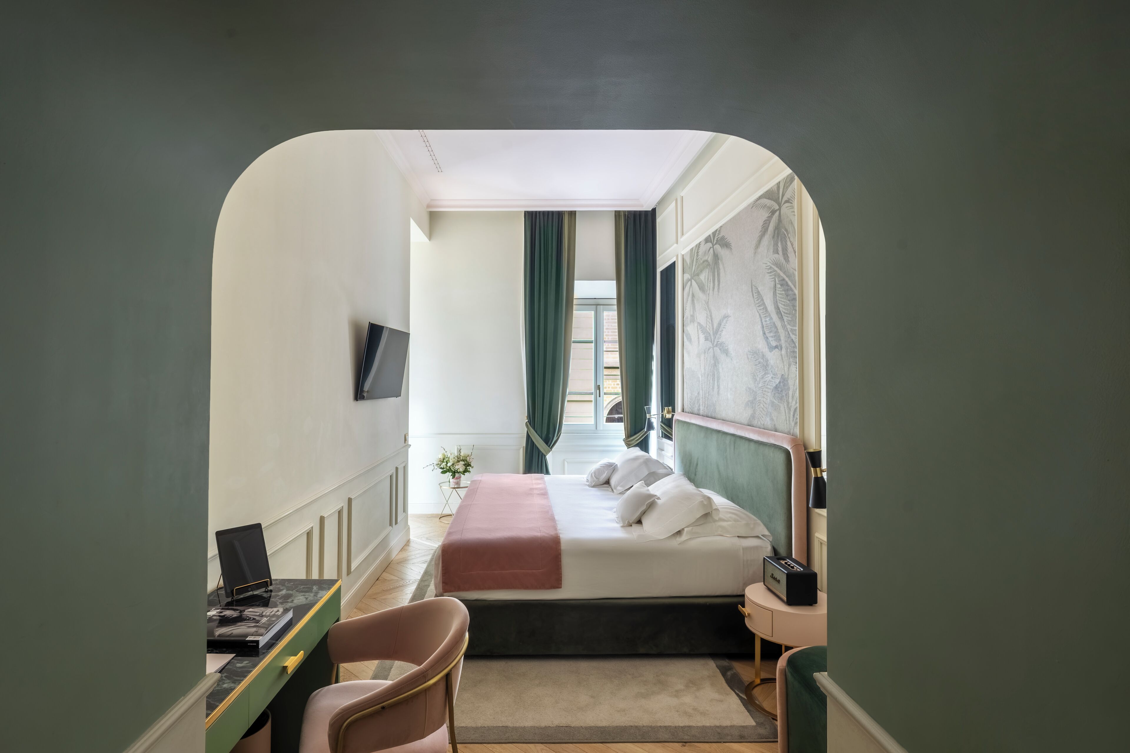 Une chambre d’hôtel avec un lit, un bureau avec un ordinateur, une chaise, une table de chevet avec une lampe, et une vue sur l’extérieur, visible à travers l’encadrement d’une porte.